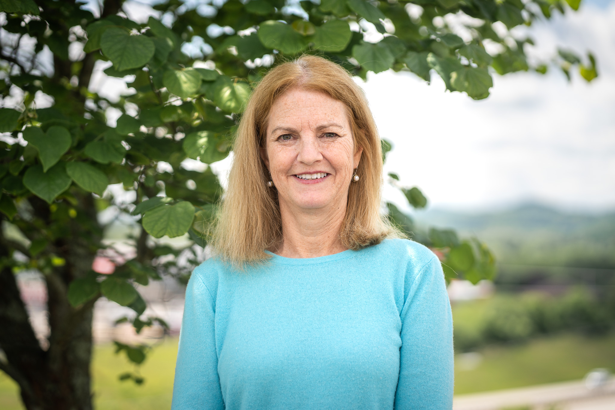Associate Provost, Nancy Parks, is pictured smiling while wearing a light blue shirt. She stands in front of a tree with mountains in the background.