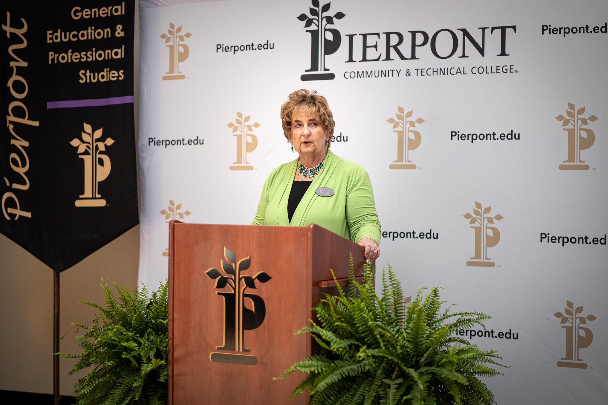 Dr. Kathleen Nelson stands behind a wooden podium adorned with the Pierpont logo, delivering a speech.