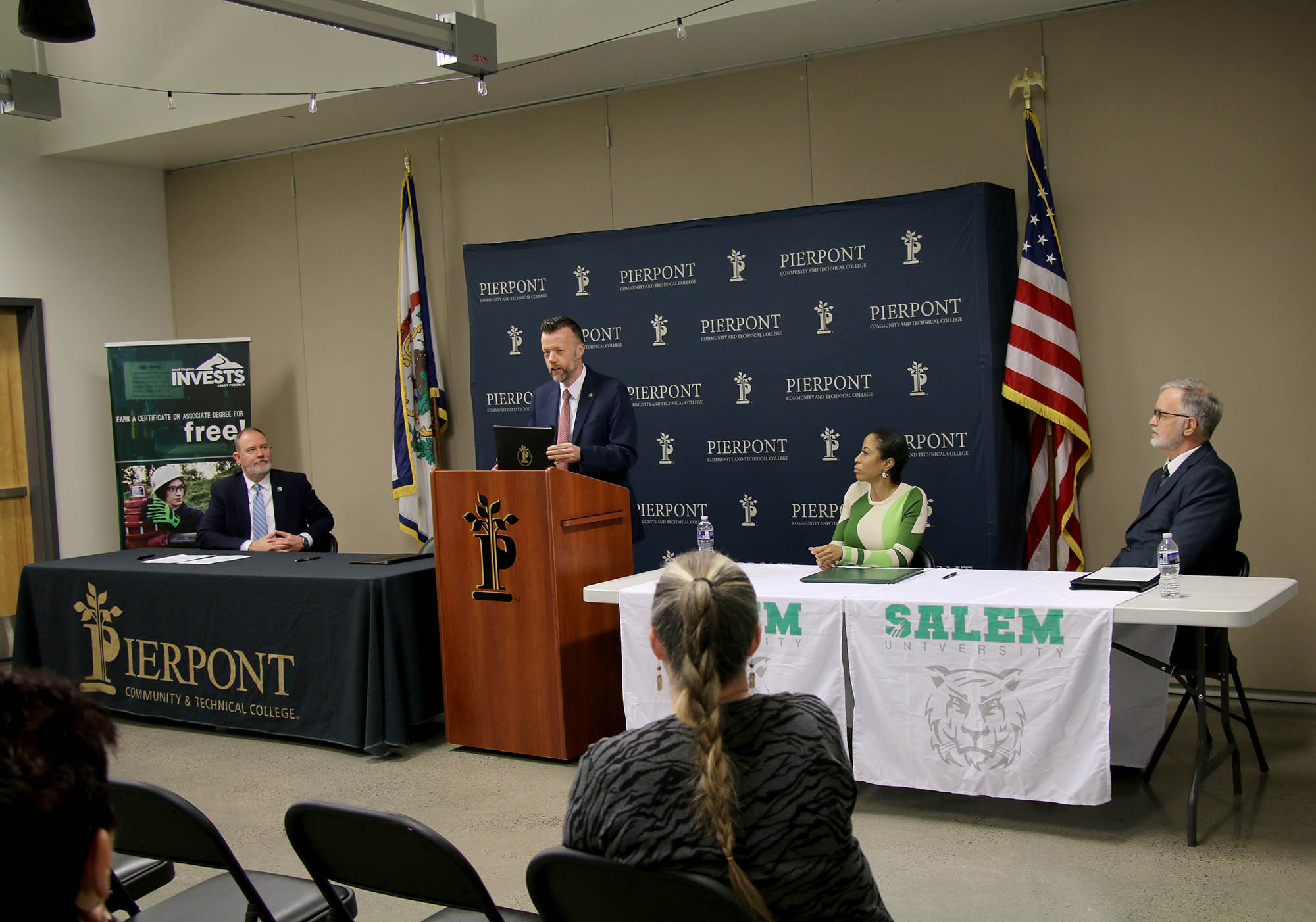 A speaker stands at a podium with Pierpont branding, addressing an audience in a conference-style room. Three individuals sit at tables on either side — one at a Pierpont‑branded table and two at a Salem‑branded table — during the signing event. An American flag and WV state flag are visible in the background.