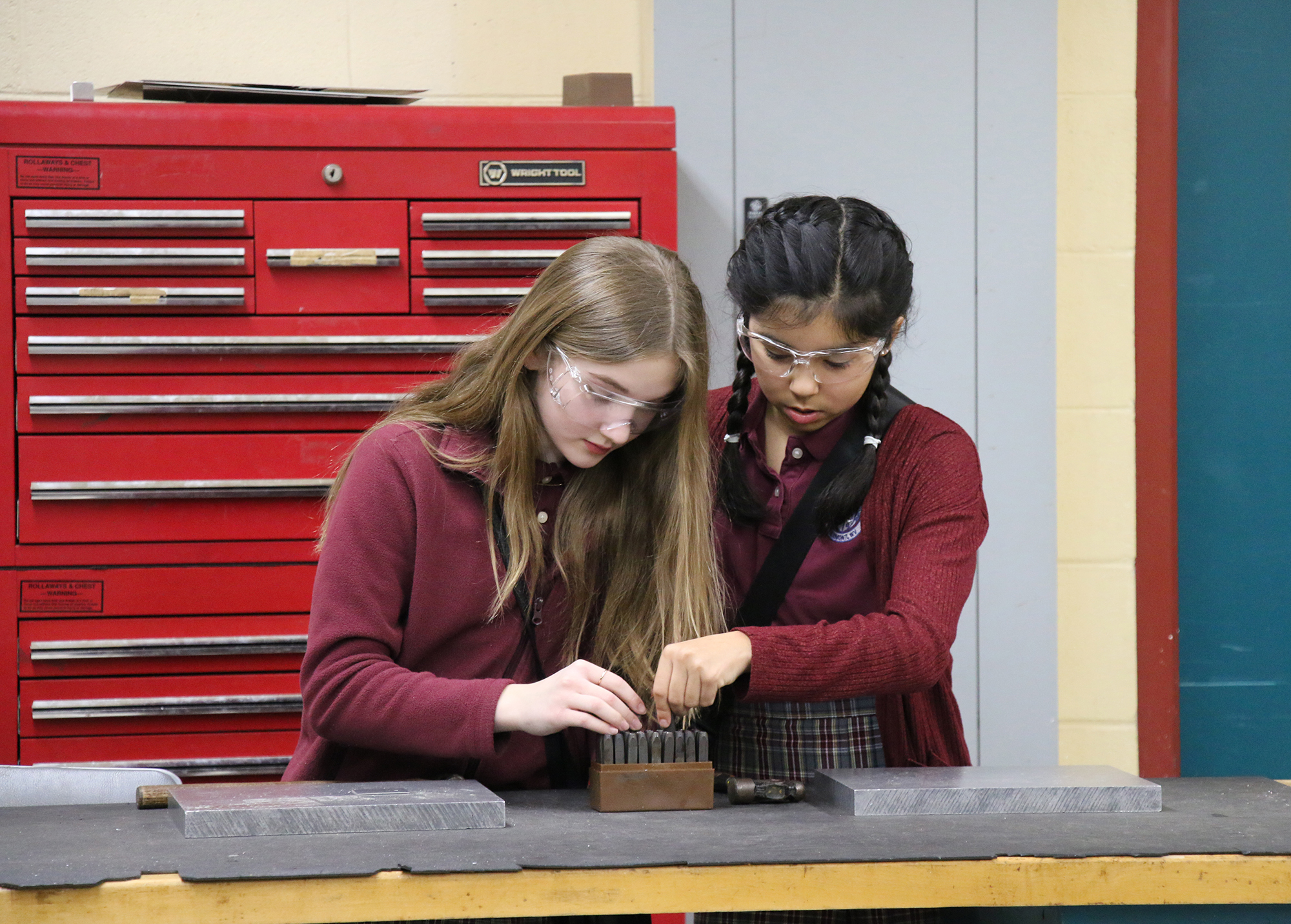 Two students stand at a workbench selecting metal stamps and working on a sheet‑metal project inside an aviation lab classroom.