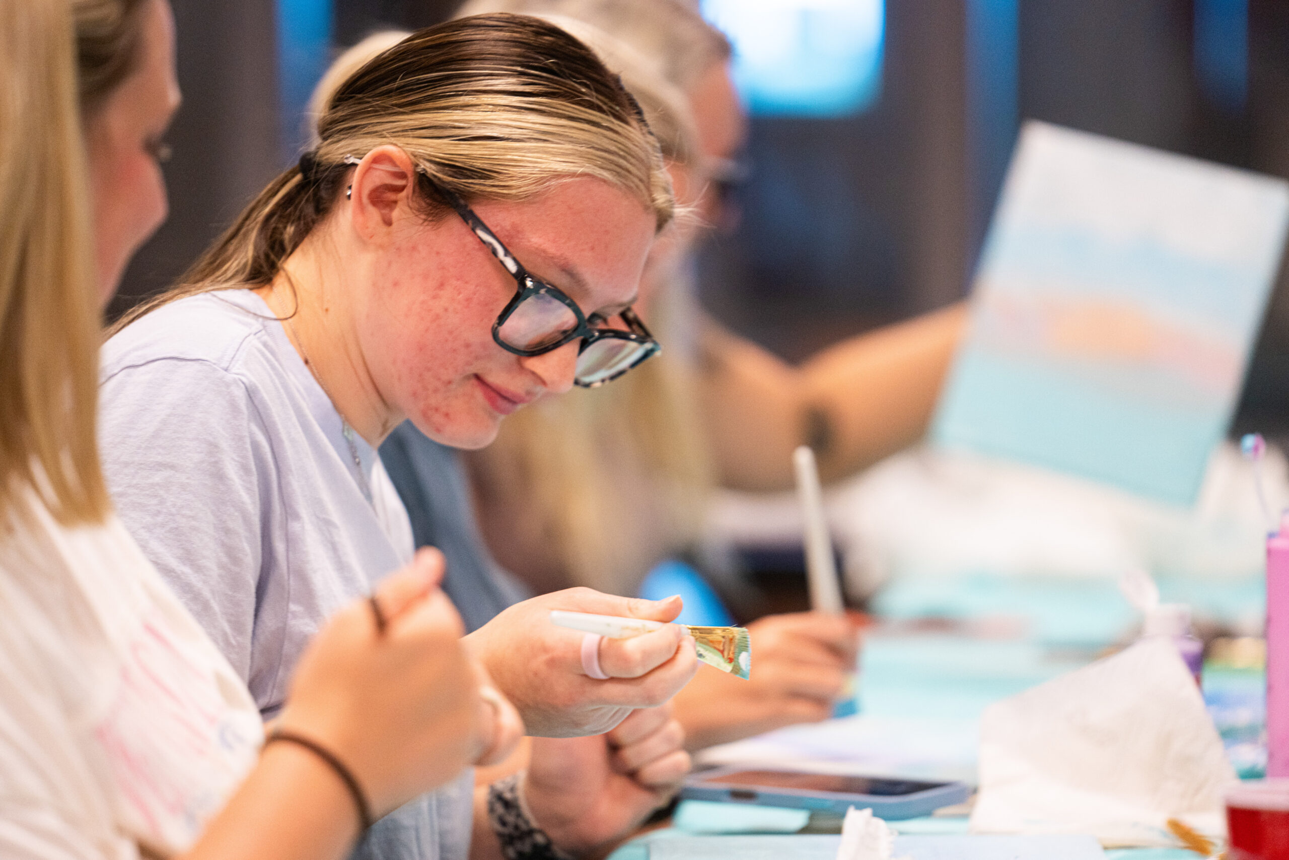 A person with glasses and blonde hair with streaks of color is painting with a brush and a small canvas in front of them. Other people are visible in the background, also engaged in painting.